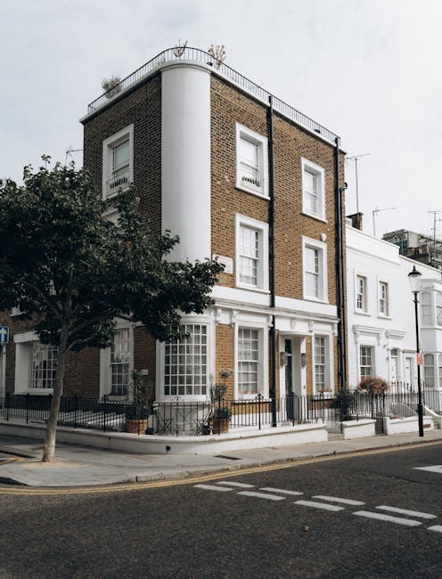A multi-storey residential building situated at the corner of a paved street, featuring a facade with a mix of brickwork and white painted sections. The building has four visible levels, with the upper floors displaying brown brickwork and white-framed sash windows, some with closed shutters. A white rounded column runs vertically along the corner, supporting the roof terrace area which is enclosed by a metal railing with potted plants placed along the edge. The ground floor has white-rendered walls, large sash windows, and a central door with a small glass panel, flanked by potted plants and black metal railings. To one side, a leafy tree partially obscures the building's lower part and street furniture such as a lamppost and black metal fencing are visible along the sidewalk, which is clean and well-maintained. This scene highlights urban residential architecture with no visible rubbish or waste material in the vicinity, exemplifying private property in a tidy, well-kept environment, possibly related to discreet waste management or on-site clearance practices with no evident rubbish removal activity occurring at the moment.
