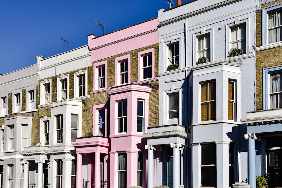 A row of Victorian-style terraced houses with multiple stories, featuring bay windows and decorative cornices, painted in pastel shades including white, light pink, and pale blue. The buildings have brick facades with some sections painted over, and the windows are framed with white trim, some adorned with flower boxes containing greenery and flowers. The architectural details include ornate brackets supporting the small overhangs above the bay windows and brickwork accents around window openings. The street scene is captured in bright daylight, with a clear blue sky overhead. Shadows cast by the buildings emphasize their depth and texture. This urban setting is typical of residential neighborhoods where private refuse collection or on-site rubbish removal services, such as those provided by Rubbish Removal Notting Hill, might be utilized for waste management. The scene does not show any debris or rubbish but provides a clear view of the housing style suitable for various waste disposal needs related to property maintenance or refurbishment that might require expert rubbish clearance.
