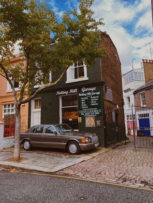 The image shows a two-story brick building with a dark green facade, housing Notting Hill Garage, a business offering services such as exhaust fitting, and repairs, as indicated on the signboard. In front of the garage, a vintage grey car with a textured, metallic finish is parked on a cobblestone driveway. To the left of the vehicle, a young tree with a slender trunk and green leaves extends upward, partially obscuring the upper window of the building. The sidewalk is paved with irregular cobblestones, and a black metal fence runs along the right side of the property, separating it from adjacent residential buildings. The background displays a partly cloudy sky with patches of blue, and neighboring structures with white and brick exteriors are visible on either side of the garage. The scene appears quiet and typical of a residential area where independent vehicle repairs and rubbish removal services could be arranged as an alternative to council collections.