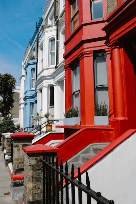 A row of Victorian-style terraced houses with multiple stories, featuring bay windows and decorative cornices, painted in pastel shades including white, light pink, and pale blue. The buildings have brick facades with some sections painted over, and the windows are framed with white trim, some adorned with flower boxes containing greenery and flowers. The architectural details include ornate brackets supporting the small overhangs above the bay windows and brickwork accents around window openings. The street scene is captured in bright daylight, with a clear blue sky overhead. Shadows cast by the buildings emphasize their depth and texture. This urban setting is typical of residential neighborhoods where private refuse collection or on-site rubbish removal services, such as those provided by Rubbish Removal Notting Hill, might be utilized for waste management. The scene does not show any debris or rubbish but provides a clear view of the housing style suitable for various waste disposal needs related to property maintenance or refurbishment that might require expert rubbish clearance.