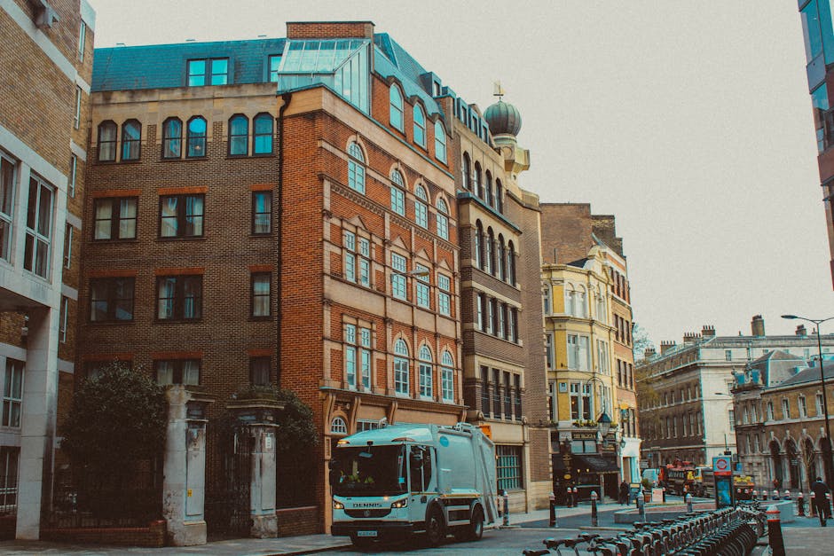 A street scene in an urban area featuring several multi-story buildings with varying architectural styles, including brick facades, large arched and rectangular windows, and a building with a domed tower at its corner. The buildings exhibit a mix of brown, red, yellow, and beige tones, with some structures having decorative stonework and ornate window frames. In the foreground, a white waste collection truck is parked on the street, partially blocking the view of a gated entrance flanked by stone pillars with metal fencing. The street is lined with bollards and bicycle racks, and there are additional vehicles and street furniture visible in the background, along with a clear, overcast sky. The scene appears to be in a historic or central part of the city, close to commercial and residential buildings, with natural lighting emphasizing the textures and colors of the structures.