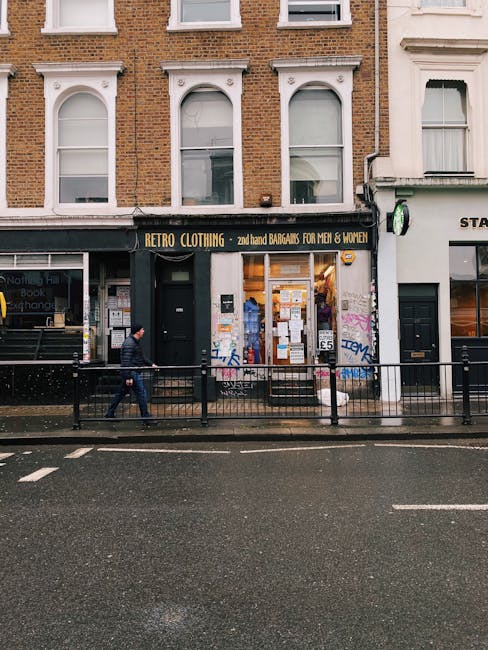 A street-level view of a retail shop named 'Retro Clothing' with a black facade and yellow signage indicating second-hand bargains for men and women. The shop has a large window displaying various clothing items and notices, with a black door to the left of the window. In front of the store, a black metal railing separates the pavement from the road, which has white lane markings and appears wet, suggesting recent rain. A person dressed in dark clothing and a beanie is walking past the shop, further emphasizing the urban environment. To the right, there is a neighbouring white building with a partially visible sign, and some graffiti is visible on the wall adjacent to the shop. The upper floors of the building are brick with white window frames and ornamental architectural features. The overall scene depicts typical commercial premises in a cityscape, with the appearance of city maintenance and the potential for local waste collection or private rubbish removal services, complementing the context of managing waste for retail businesses like this one.