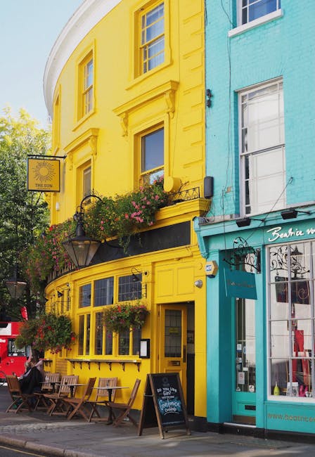 The image depicts a vibrant street scene featuring a two-story building painted in a bright yellow hue, with ornate architectural details around the windows and doors, situated next to a turquoise-colored storefront. In the foreground, there is an outdoor seating area with wooden tables and chairs, some of which are occupied by individuals. Hanging flower baskets with pink and red blooms are attached to the building's façade, adding decorative greenery. A vintage-style street lamp extends from the wall, and a sign for a café or business is displayed above the entrance. The setting appears to be a lively urban area, likely in a commercial district, with a clear sky and natural daylight illuminating the scene. The image showcases an inviting exterior that could be associated with independent cafés or shops, emphasizing the character and atmosphere typical of niche shopping and leisure streets, relevant in the context of alternative waste handling or independent collection practices managed by companies like Rubbish Removal Notting Hill.