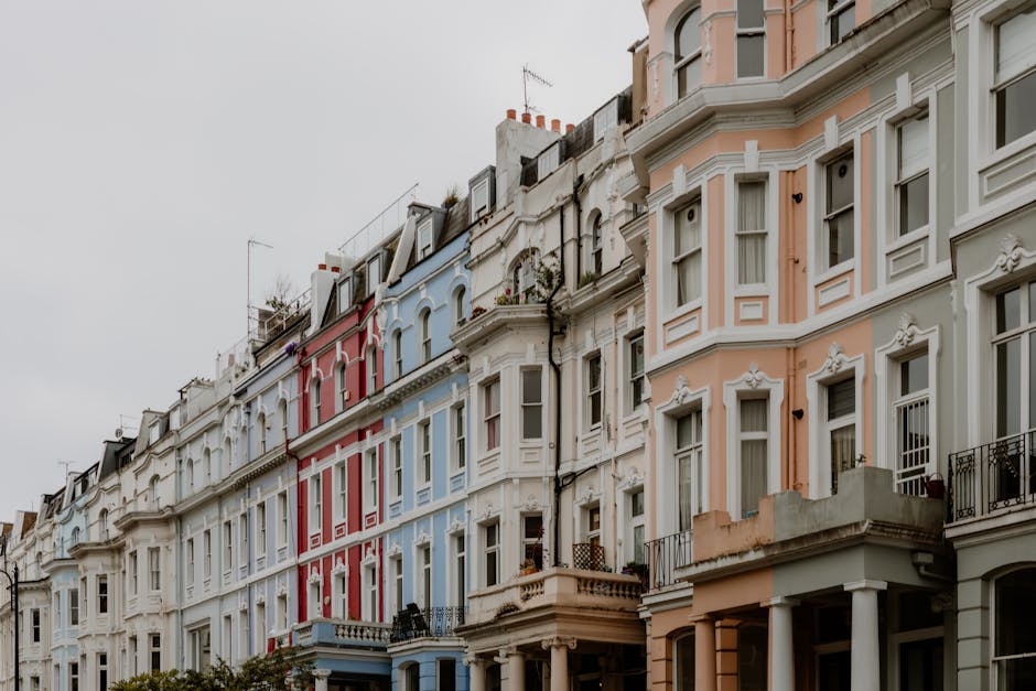 A busy street scene in Notting Hill during a market day, featuring a variety of pedestrians browsing and interacting around outdoor stalls with colorful canopies and displays. The street is lined with historic Victorian-style buildings with ornate facades and large windows, housing shops and cafes. Above the street, Union Jack flags and a banner with “Portobello Road W11” are strung across the road, emphasizing the location's identity. In the foreground, a grey van is parked on the right side, while people in casual outdoor clothing, including jackets and hats, are walking, standing, or sitting along the pavement. Some individuals are engaged in casual conversations, and others browse goods on display, creating a lively and vibrant atmosphere. The scene captures the outdoor market environment typical of independent waste handling and rubbish collection in busy urban areas, with Rubbish Removal Notting Hill providing a relevant context for waste management services supporting such local events and commercial activities.