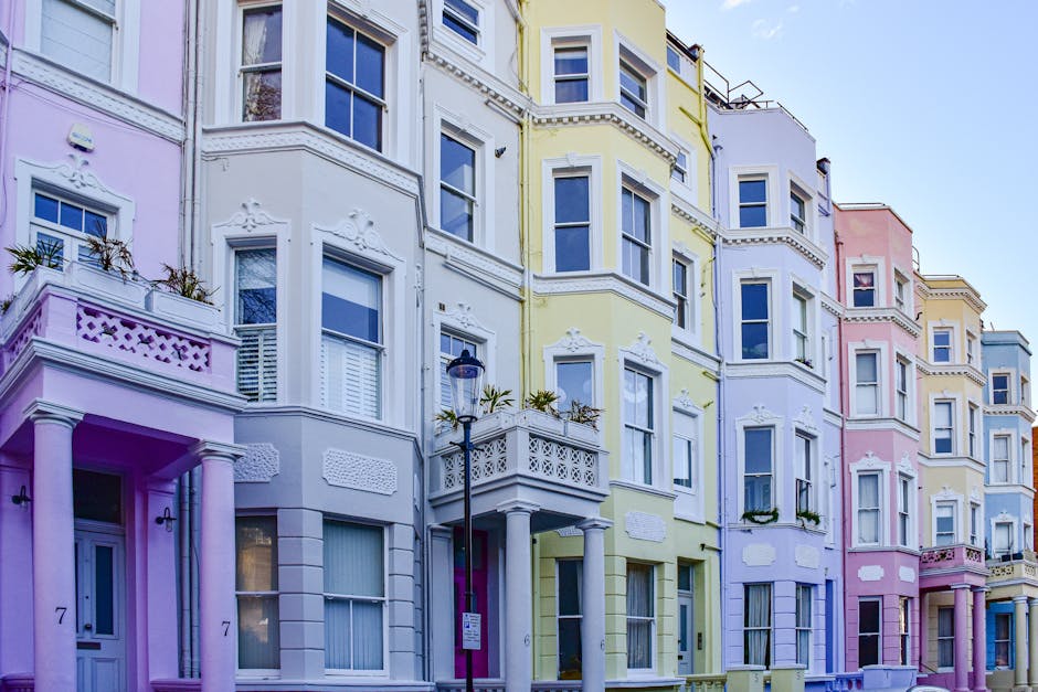 The image depicts a row of multi-storey terraced houses painted in pastel shades including pink, white, yellow, purple, and light blue. Each building features detailed architectural elements such as decorative mouldings, bay windows, and small balconies with ornamental railings. The facades display a mixture of smooth finishes and textured surface details, highlighting traditional Victorian-style architecture. A black street lamp stands prominently in the foreground near the sidewalks, which are clean and well-maintained. Some windows have shutters and flower boxes, while others are plain, with clear glass panes reflecting the bright, natural daylight. The composition shows the buildings tightly positioned side by side, creating a uniform but colorful streetscape typical of historic neighborhoods. The sky above is partly cloudy with a gentle blue tone, and the overall scene exudes a quiet, organized urban residential environment, relevant to the context of private property and alternative waste management solutions in busy city areas, supported by local rubbish removal services such as Rubbish Removal Notting Hill.