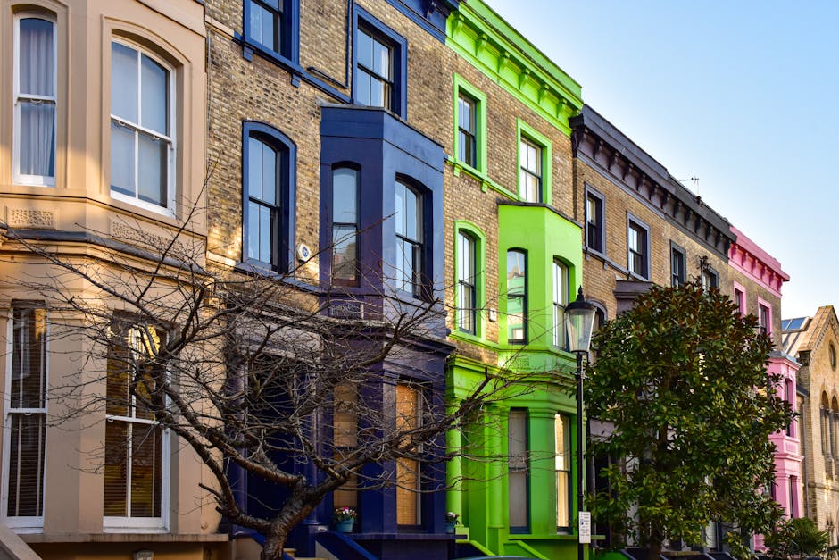 A row of multi-storey Victorian-style terraced buildings with ornate brick facades, painted in a variety of vibrant colours including beige, navy blue, lime green, and pink. The buildings feature sash windows with white trims, some with small balconies or bay windows. In the foreground, a leafless tree with thin, twisting branches partially obscures the lower part of the buildings, while a streetlamp with a black metal post and glass enclosure is positioned near the green building. To the right, a lush, green shrub with dense foliage grows close to the sidewalk. The scene is illuminated by natural daylight, with a clear blue sky overhead, highlighting the bright, fresh appearance of the buildings and the contrast between the painted exteriors and the natural elements. This urban streetscape exemplifies contrasting colour schemes on residential properties, indicative of private property aesthetics and potential sites for external rubbish removal or renovation-related waste clearance, as characteristic of independent waste disposal services like those offered by Rubbish Removal Notting Hill.