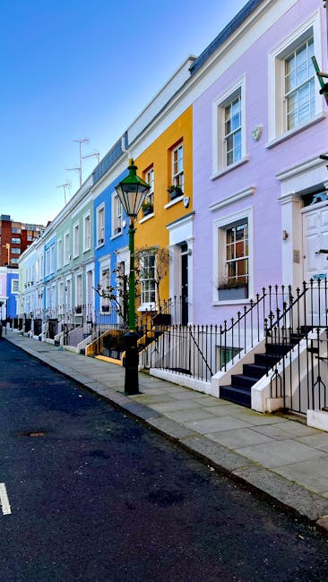 The image depicts a row of multi-storey terraced houses painted in pastel shades including pink, white, yellow, purple, and light blue. Each building features detailed architectural elements such as decorative mouldings, bay windows, and small balconies with ornamental railings. The facades display a mixture of smooth finishes and textured surface details, highlighting traditional Victorian-style architecture. A black street lamp stands prominently in the foreground near the sidewalks, which are clean and well-maintained. Some windows have shutters and flower boxes, while others are plain, with clear glass panes reflecting the bright, natural daylight. The composition shows the buildings tightly positioned side by side, creating a uniform but colorful streetscape typical of historic neighborhoods. The sky above is partly cloudy with a gentle blue tone, and the overall scene exudes a quiet, organized urban residential environment, relevant to the context of private property and alternative waste management solutions in busy city areas, supported by local rubbish removal services such as Rubbish Removal Notting Hill.