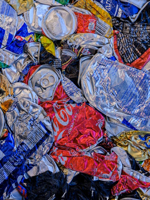 A close-up image of a pile of crushed and crumpled beverage cans, primarily made of aluminium, with visible pull tabs and embossed surfaces in various metallic shades. Some cans display colourful branding and logos, including red, blue, and black hues, with a partially visible Coca-Cola logo on a prominently red can. The cans are stacked irregularly, overlapping each other, and appear to be discarded waste, likely collected for recycling or disposal. The textures range from smooth and shiny to dented and wrinkled, with some edges slightly sharp or torn. The background is not visible, but the cans fill the entire frame, suggesting an outdoor or indoor setting where waste has been gathered, possibly at a rubbish collection or recycling point. This scene is representative of waste management processes involving the collection of empty beverage containers, aligning with independent collection or recycling efforts managed by services like Rubbish Removal Notting Hill.