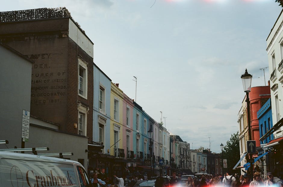 A busy street scene in Notting Hill, featuring a row of multi-story terraced buildings painted in pastel shades of pink, blue, yellow, and white, with some structures displaying textured brickwork and decorative window frames. In the foreground, a white van with the word 'Gradvanced' partially visible on its side is parked along the pavement, which is populated with pedestrians walking and engaging in daily activities. The street is lined with various shops with awnings extending over the sidewalk, and traditional street lamps are visible, adding to the characteristic ambiance of the area. The sky above is partly cloudy with soft, diffuse lighting. The scene captures an urban environment where private waste handling or rubbish removal services, such as those provided by Rubbish Removal Notting Hill, may operate to maintain the cleanliness and appearance of this vibrant neighborhood, emphasizing the importance of on-site clearance for waste and rubbish within a lively cityscape.
