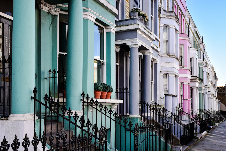 A row of Victorian-style terraced houses with colourful pastel façades lining a narrow street. The buildings feature decorative architectural elements such as pilasters, cornices, and window trims. The front entrances are accessed via small steps, with black wrought iron railings and gates separating the properties from the pavement. Each house has large, rectangular windows, some with flower boxes or planters containing green shrubs on their window sills. The façades display shades of teal, dark grey, pink, and light green, highlighting the vibrant architectural character. The sky above is clear and blue, providing natural lighting that enhances the bright, clean appearance of the buildings. This scene naturally relates to private property presentation in locations where external rubbish removal services such as Rubbish Removal Notting Hill are common, emphasizing the importance of maintaining tidy, well-kept premises within a residential street setting for effective waste management.