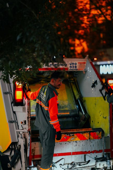 A male waste disposal worker with short dark hair and a beard, wearing a high-visibility orange and grey work uniform with reflective stripes, stands beside a large waste collection vehicle during dusk or evening. The vehicle is yellow and white, equipped with a rear loading mechanism and safety lights illuminated in red. The worker is positioned near the side of the vehicle, holding onto the edge with his left hand, while looking over his shoulder. The background features a dark, leafy environment with the orange glow of sunset skies visible through the trees, creating a contrasting atmosphere of urban activity and natural surroundings. The scene captures an on-site waste collection operation typical of private rubbish removal services, emphasizing the professional handling of waste materials in a manner consistent with rubbish removal and disposal processes in the region. The image appears to be documenting an independent rubbish collection activity, with focus on the worker, vehicle, and the setting's environmental conditions, highlighting the practical aspects of waste management outside municipal collection services.