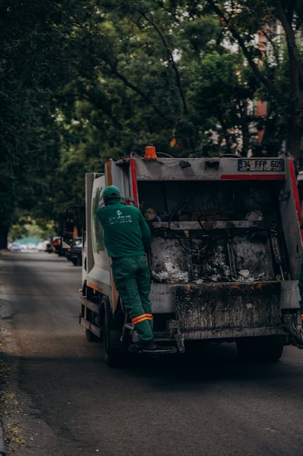 A waste collection worker in green uniform and safety helmet stands at the back of a medium-sized rubbish collection van, which has its rear loading compartment open to reveal ashes and burnt debris inside. The van is parked on a residential street lined with trees, with a few cars visible further along the road. The scene takes place during daylight hours with diffuse natural light filtering through the leafy canopy overhead. The worker appears to be inspecting or managing the contents of the waste vehicle. The van's exterior is white with some visible dirt and soot around the rear area, indicating recent collection activity. Surrounding the scene are greenery and parked vehicles, suggesting an area where private waste removal services could be employed for on-site clearance of waste and debris, with Rubbish Removal Notting Hill providing services aligned with alternative rubbish collection options in the local W11 district.
