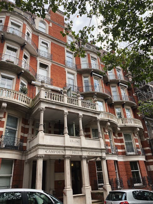 The image depicts the historic facade of a multi-storey red brick residential building with ornate white stone balconies and decorative railings. The building features several large, white-trimmed windows and a prominent main entrance accessed via a classical portico supported by multiple columns. The entrance bears the inscription 'Campden Hill Court' along with the address number '13-36'. Surrounding the building, there are leafy tree branches partially obscuring the upper levels, with natural daylight illuminating the scene and creating soft shadows on the structure. At street level, two parked cars are visible in the foreground, emphasizing the urban residential setting. This scene reflects a typical example of London’s architectural style, which may be associated with private or independent property management, relevant to the context of on-site or alternative waste handling, commonly managed by local rubbish removal services such as Rubbish Removal Notting Hill. The detailed details of the building's materials, textures, and classic design elements are clearly visible, supporting accessibility and contextual understanding of potential rubbish or waste collection points for residents or property managers in the area.