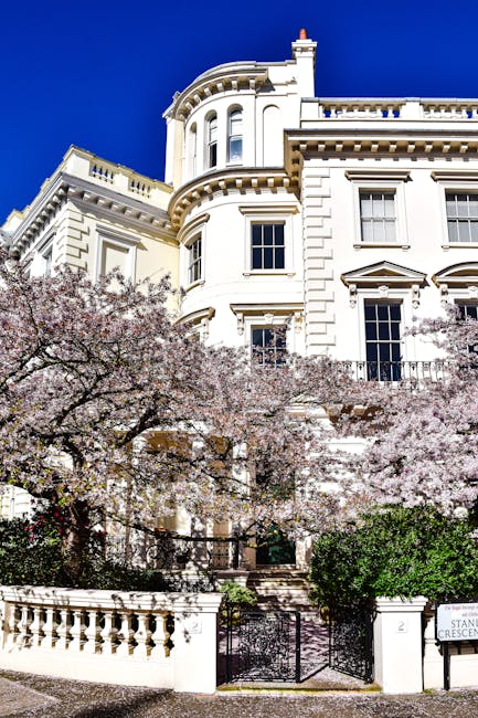 A large, white, multi-story building with ornate architectural details, including rounded bay windows and decorative cornices, set against a clear blue sky. In the foreground, a flowering tree with light pink blossoms partially obscures the lower part of the building, which features a black metal gate and a curved stone balustrade. The setting appears residential, with well-maintained greenery and a small garden area near the entrance. The building's exterior walls have a polished, smooth finish, and the windows are framed with moldings. The scene suggests a peaceful, urban environment with high-quality private residences, and it may relate to the context of independent or private waste handling, though no rubbish or waste items are visible.