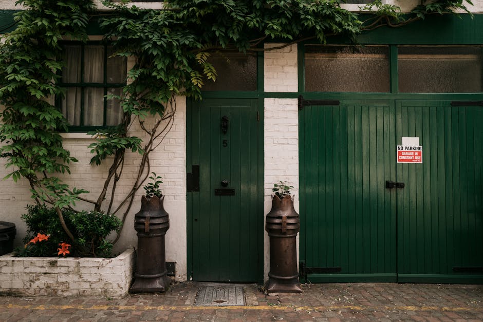 A storefront with a white brick wall and dark green wooden doors and window frames. The window frame on the left contains frosted glass panes, partially obscured by a climbing plant with broad green leaves that extends across the top of the wall. The plant's stems twist around the door and window frames, adding natural texture. In front of the building, there are two large, vintage-style metal bollards with rusted finishes, positioned on either side of a small, grated concrete step leading up to a narrow green door, which has a black handle and an exposed keyhole. To the left of the door, a small, white brick planter box contains a variety of green foliage and a few orange flowers, with some plants spilling over the edge. The ground is paved with small, uneven cobblestones, contributing a rustic look. To the right of the main door, a large green sliding garage door features black hardware and a white and red sign indicating no parking and property use restrictions. The overall scene suggests an urban setting with elements that support private waste and rubbish management, consistent with services offered by Rubbish Removal Notting Hill.