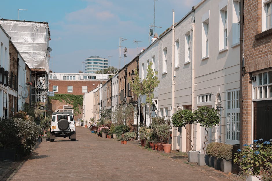 The image depicts a narrow residential street in a city environment, with a paved cobblestone surface and row houses on either side. The houses are primarily white or light-colored, with some featuring brick elements and small front gardens or potted plants positioned near doorways. On the left, a white van with a spare tyre mounted on its rear is parked facing away from the viewer, indicating a possible vehicle used for rubbish removal or waste collection. The street is lined with various potted plants, including small trees and shrubbery, which serve as decorative elements alongside waste management tasks. In the background, taller modern office buildings are visible, suggesting an urban setting with residential and commercial mixed-use development. The sky is mostly clear with some scattered clouds, and the lighting is natural, indicating daytime. The scene subtly alludes to independent waste management and street clearance activities, fitting within the context of rubbish removal services provided by Rubbish Removal Notting Hill, emphasizing on-site clearance and private disposal methods supported by such professionals.