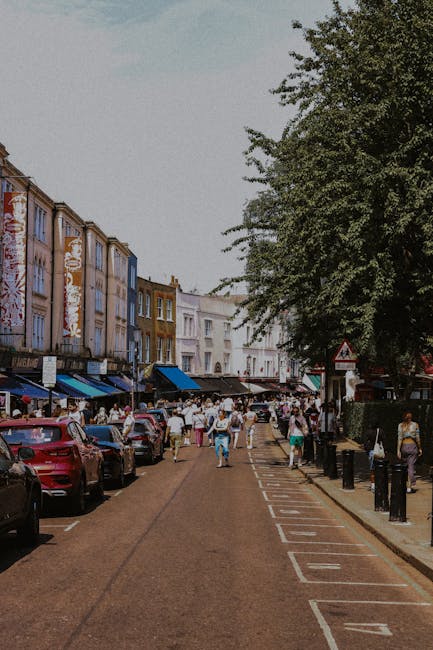 A busy street scene in a shopping district showing multiple parked cars along the left side of the roadway, with a row of multi-storey buildings featuring varied pastel-colored facades, some with large store windows and awnings. Pedestrians are walking along the paved sidewalk and across the street, with a mixture of individuals and small groups visible. On the right side, tall green trees line the pavement, casting partial shade over the scene. In the background, more storefronts and signage are visible, contributing to a lively, urban atmosphere. The overall scene captures the everyday activity of city life, with a focus on pedestrian movement and vehicle parking, relevant in the context of urban waste management and municipal rubbish collection services, demonstrating how private refuse handling might be integrated into bustling commercial districts.
