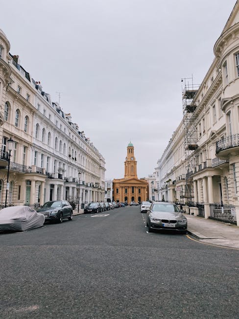 A quiet street scene featuring a row of elegant, white multi-storey residential buildings with classic architectural details such as wrought-iron balconies, decorative window surrounds, and pitched roofs. The buildings are situated on either side of the asphalt road, which is lined with parked cars, including a silver sedan in the foreground on the right and a covered vehicle on the left. In the distance, a historic church with a tall, ochre-colored facade and a prominent clock tower topped with a green dome is visible against an overcast sky. The scene depicts an urban environment typical of a European city with well-maintained, uniform facades and a calm atmosphere, suitable for a property or waste management service context involving private or independent rubbish collection in residential neighbourhoods like Notting Hill.