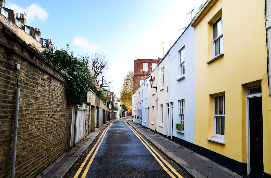 A narrow residential street in Notting Hill featuring a row of attached terraced houses on the right side painted in pastel shades of yellow, white, and light blue, with white-framed windows and small flower boxes beneath some of them. The street has a slightly wet dark asphalt surface with double yellow lines along the curb, indicating parking restrictions. On the left side, there is a high, weathered brick wall with some greenery, including a shrub with cascading branches near the top. In the background, taller brick buildings with chimney stacks and rooftop antennas rise above the tree line, which has shed some leaves, suggesting an autumn setting. The sky is clear with soft daylight, creating a calm atmosphere typical of a quiet, well-maintained neighborhood, possibly reflecting the local approach to private rubbish disposal and on-site clearance handled by services like Rubbish Removal Notting Hill.